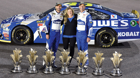 Three people stand between a race car and trophies on the ground