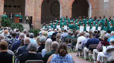 People sit in chairs facing a brick building where people in green gowns stand