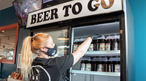 A person wearing a mask and gloves reaches into a cooling case that says Beer To Go on the top