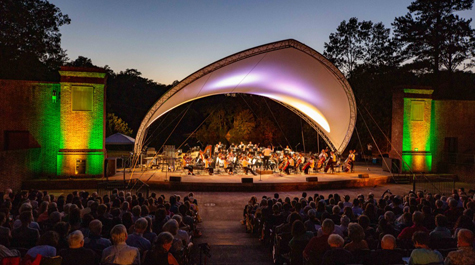 A large canopy covers an orchestra on an outdoor stage as it plays for a large crowd in the evening