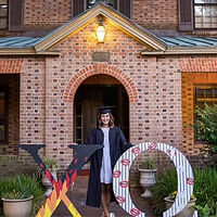 A student dressed in regalia is shown standing in front of a sorority house 