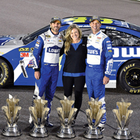 Three people stand between a race car and trophies on the ground