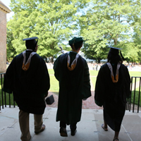 Three people in caps and gowns walk away from the camera and into an outdoor space