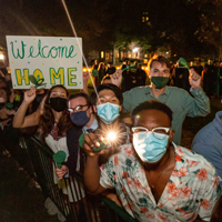 People wearing masks stand next to a fence and one holds a sign that says welcome home