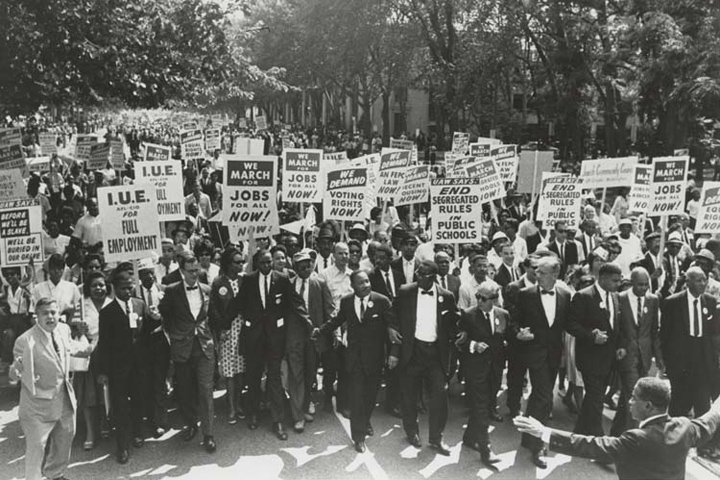A black and white photo shows a large group of people marching and holding signs about employment