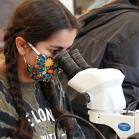 A person wearing a mask with flowers on it looks into a microscope