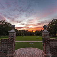 An orange and purple sunset is visible beyond two brick pillars in front of a large green space