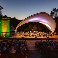 A large canopy covers an orchestra on an outdoor stage as it plays for a large crowd in the evening