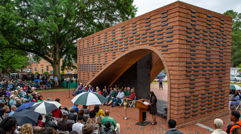 A person stands at a podium in front of a large brick structure while people sit in chairs around the space