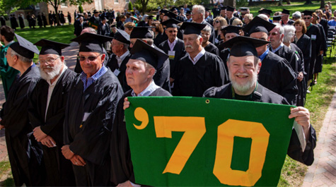 Older people wearing caps and gowns stand in a crowd and one holds a sign that says '70