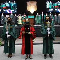 People wearing academic regalia hold maces in front of a stage with more people in regalia
