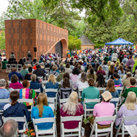 A person stands at a podium in front of a large brick structure while people sit in chairs around the space