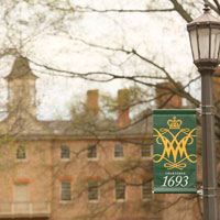 A light pole banner with the cypher and 1693 printed on it in front of a portion of the Wren Building