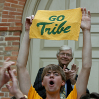 A student holds a towel that says go Tribe in front of the Wren Building with President Taylor Reveley standing in the background