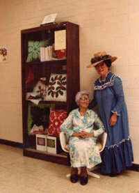 Robbins' grandmother and great-grandmother pose for a photo next to a case that displays some of their original quilting works.