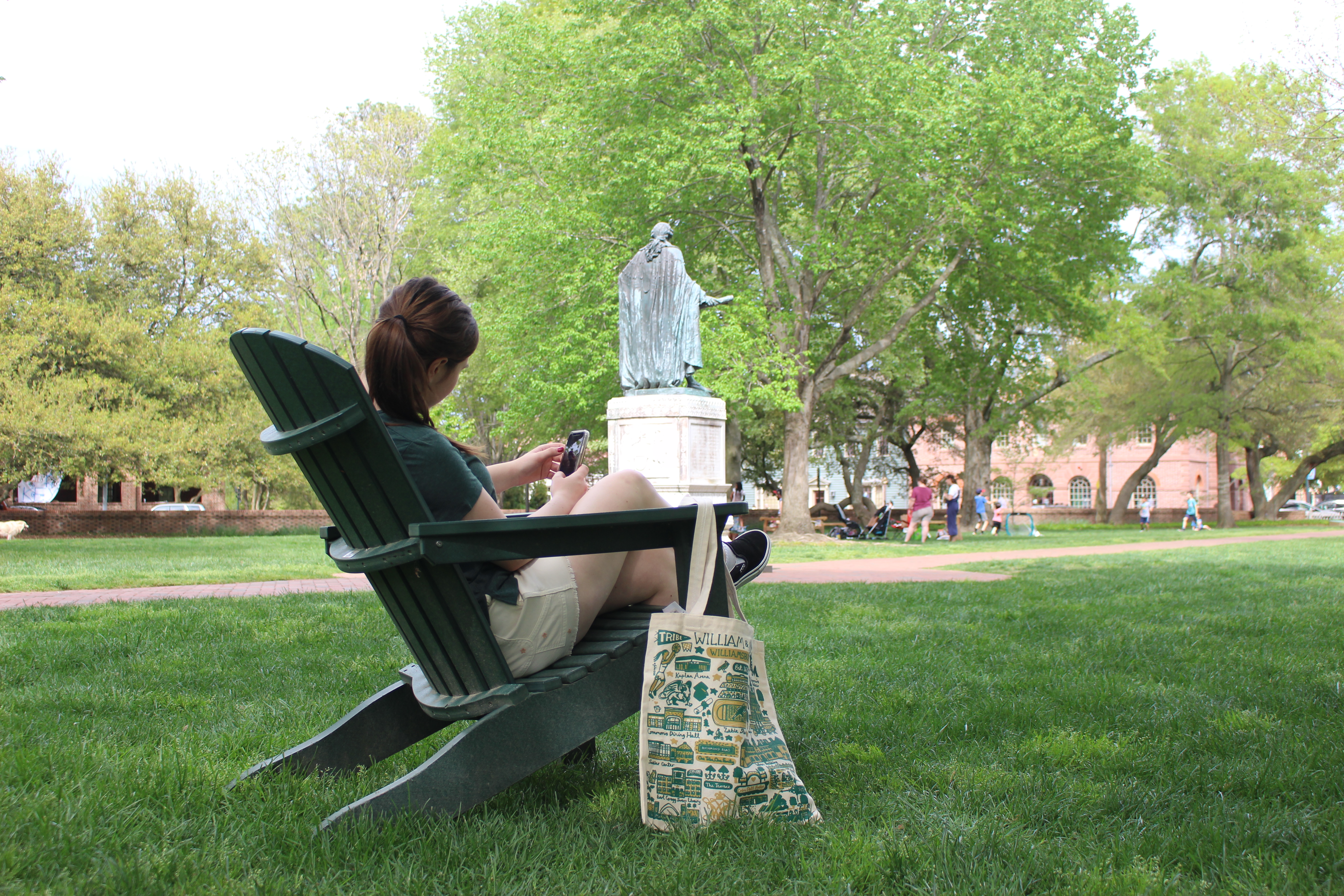 a students sits in an adirondak chair on campus. The Lord Botetout statue appears in the background and a tote bag with W&M themed illustrations sits on the ground.