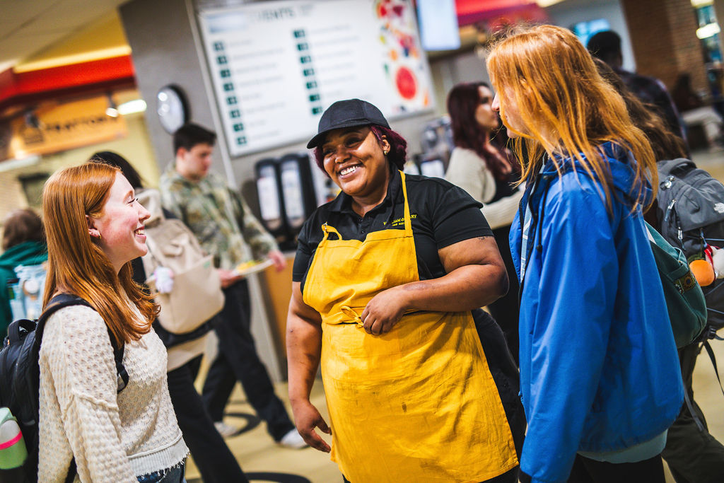 A W&M Dining employee in a golden apron smiles and laughs with two female-presenting students.