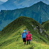 Two people walking on a grassy mountaintop with mountains in the background.