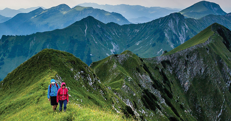 Two people walking on a grassy mountaintop with mountains in the background.