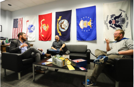 A bright office space with the U.S. and military flags hanging on the wall behind comfortable conversation chairs and couches with two students smiling and talking with Charlie Foster