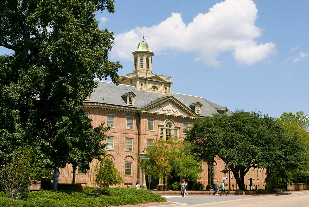 Four story colonial brick building with a large cupula surrounded by mature trees 