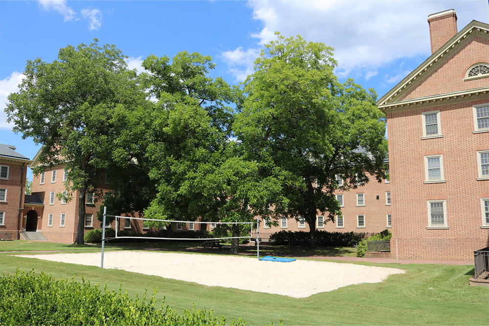 A sandy volleyball court surrounded by a brick, windowed dorm building.