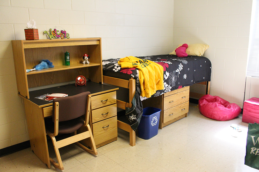 A staged dorm room shows a raised XL twin bed, a dresser under the bed, a shelf over a desk with drawers, and a chair.