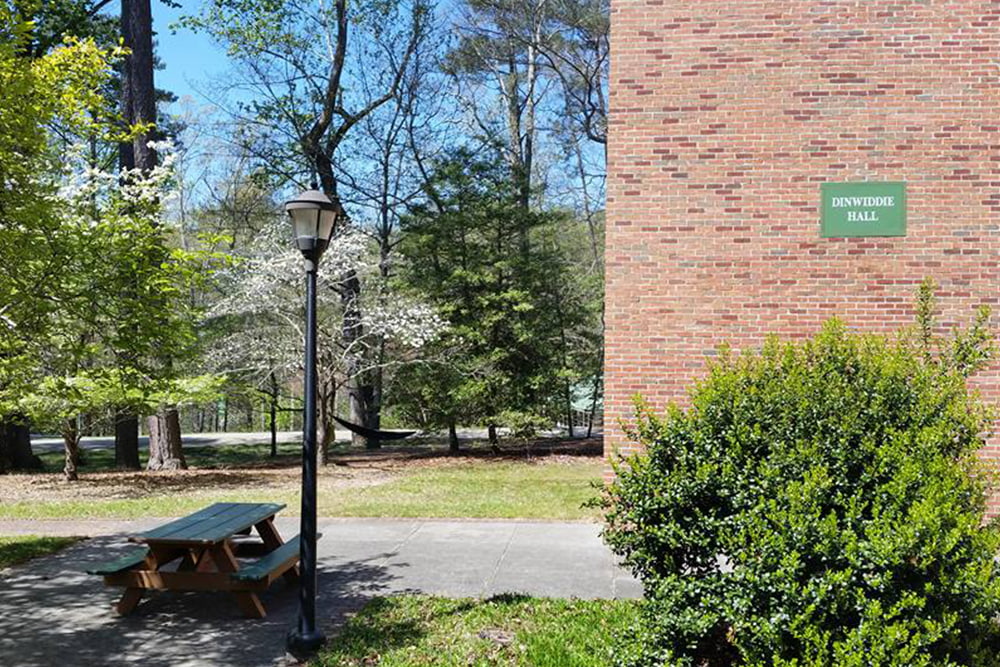 A corner of a brick dorm building sits near a paved walkway, picnic table, street lamp, and greenery.