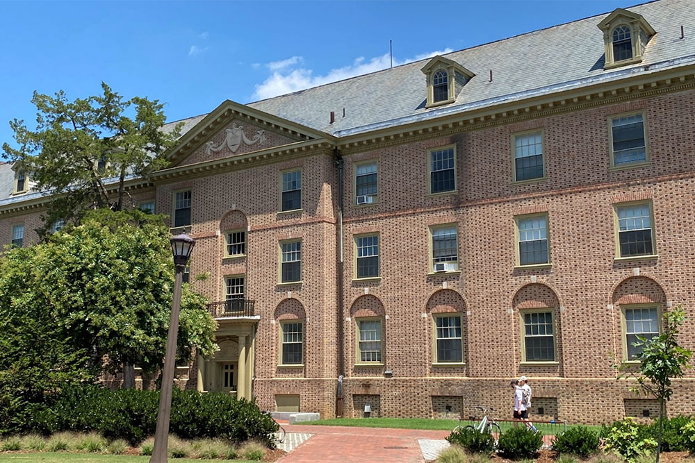 Monroe Hall, a four story brick residence hall with classic colonial architecture and dormer windows, surrounded by mature trees and landscaping