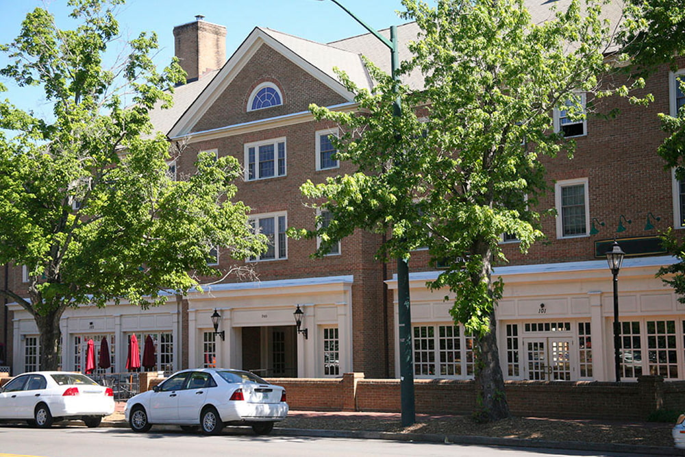 Brick dorm building with first-level shops, seated areas, and street parking next to a main road. 