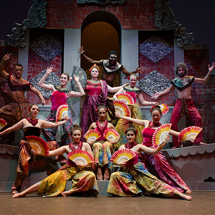 Stage performance featuring a group of dancers in colorful costumes posing with open fans against a decorative brick set.