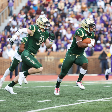 Two W&M football players in green uniforms running with the ball during a game, with a crowd in the background.