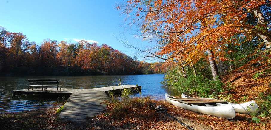 Lake Matoaka with a wooden dock and bench, surrounded by autumn trees, with canoes resting on the shore.