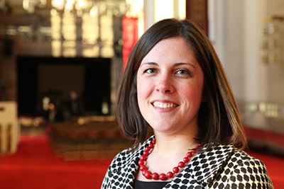 Portrait of a person with shoulder-length dark hair wearing a red necklace, standing in a room with red carpeting.