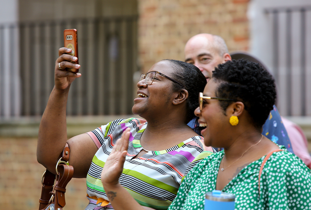 Two William & Mary employees smiling while one takes a photo with their cell phone.