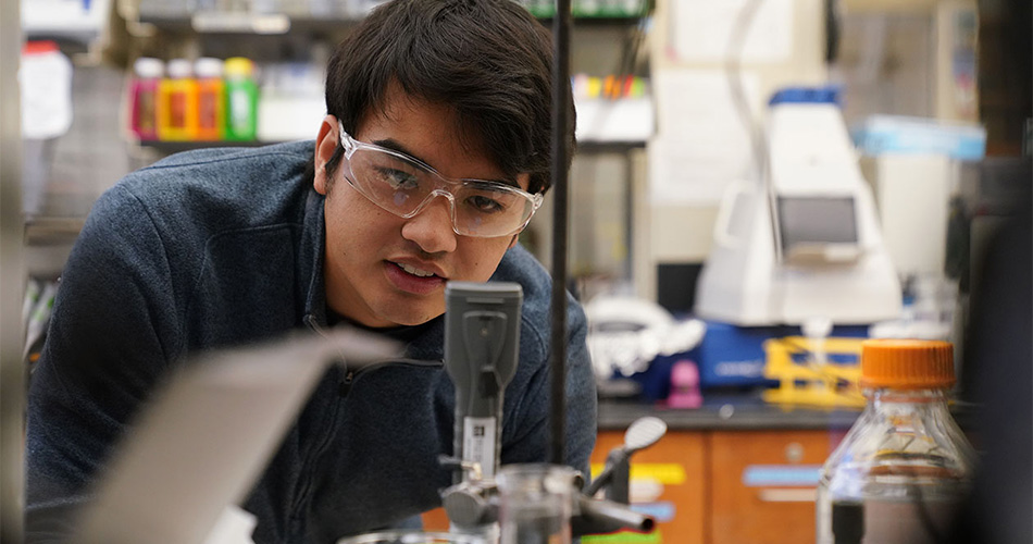A student wearing safety goggles works closely with lab equipment, focusing on a small instrument amid glassware and scientific tools.