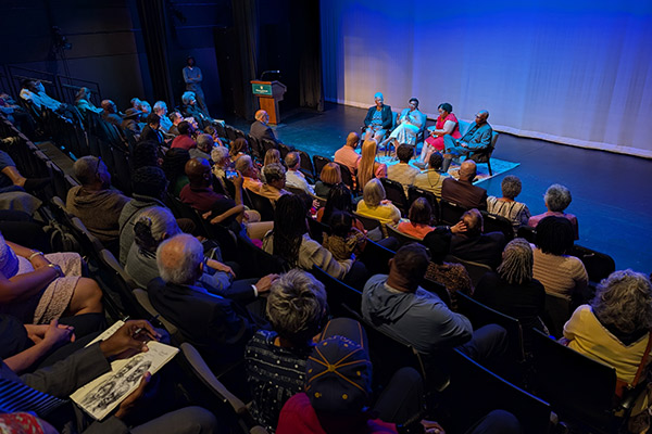 Four people seated in chairs on a blue lit stage with an audience in rows of seats watching