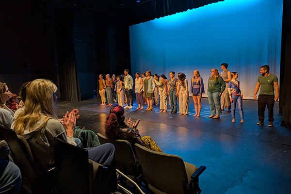 A row of student performers lined up on a blue lit stage as audience members applaud
