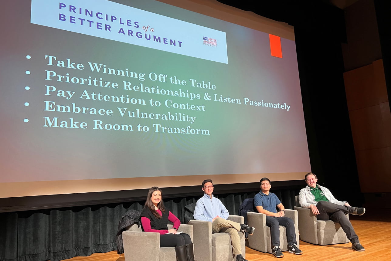 Smiling students on a stage in chairs with a slide showing the 5 principles of better arguments projected behind them