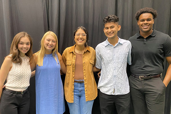 Five smiling students posed in front of a curtain background