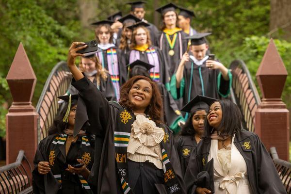 Group of graduates in caps and gowns walking across a campus bridge, smiling and taking selfies together during a graduation ceremony.