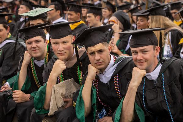 Four graduates in caps and gowns sit together at a commencement ceremony, posing with their chins resting on their hands while other graduates fill the background.