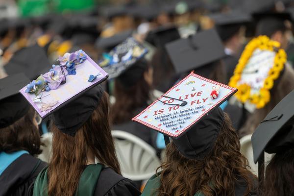 Graduates seated at a commencement ceremony wearing decorated mortarboards, including one that reads “To Live Is to Fly” and another adorned with flowers.