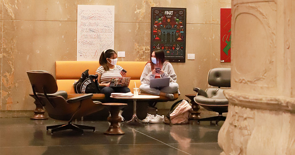 Two masked students comfortably sitting and studying together in the Botetourt Gallery on the ground floor of Swem Library