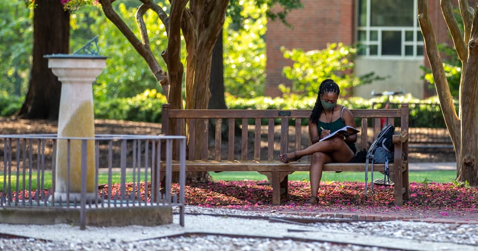A masked student sitting on a bench writing in a book near the Swem Library sundial
