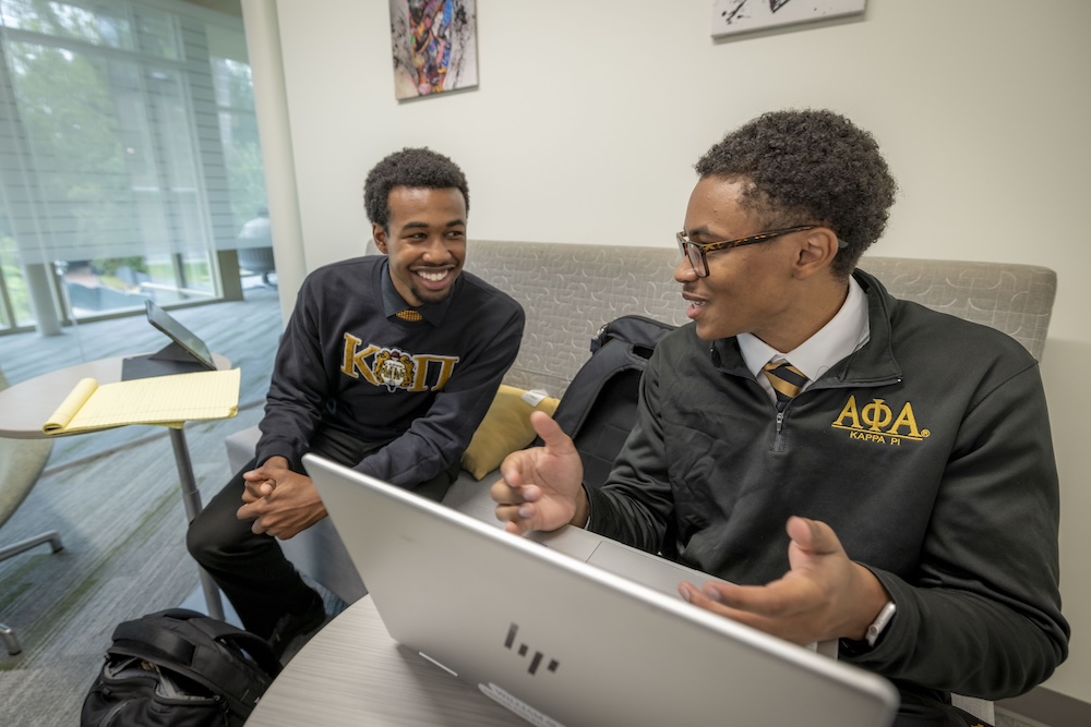 Two students seated in a lounge talking with a laptop