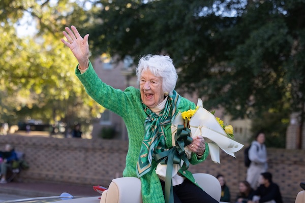Jane Batten in a green jacket and holding yellow roses waves at the crowd as part of the homecoming parade