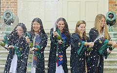 Five students in graduation robes popping bottles of sparkling in front of the Wren