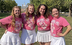 Four smiling students in pink shirts and white skirts in front of the Governor's Palace in Colonial Williamsburg