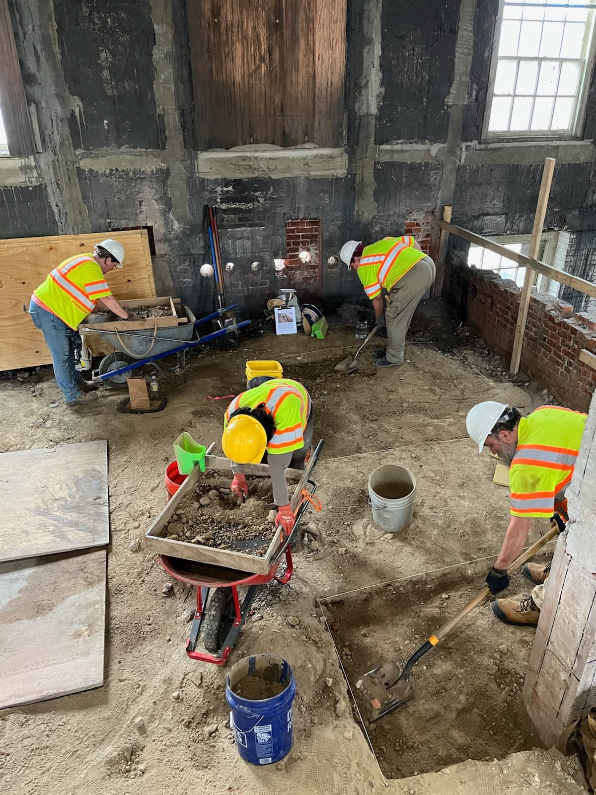 Within brick and wood walls, four archaeologists in yellow safety vests and helmets sift through the dry dirt of the cellar with shovels and screens, collecting artifacts and measuring soil levels.